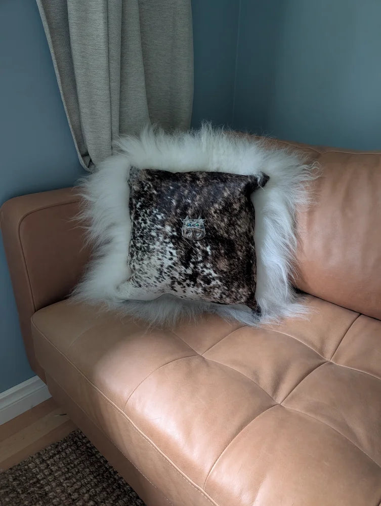 The moo cushion is a plush square pillow with a black and white speckled pattern and white faux fur trim on a tan couch