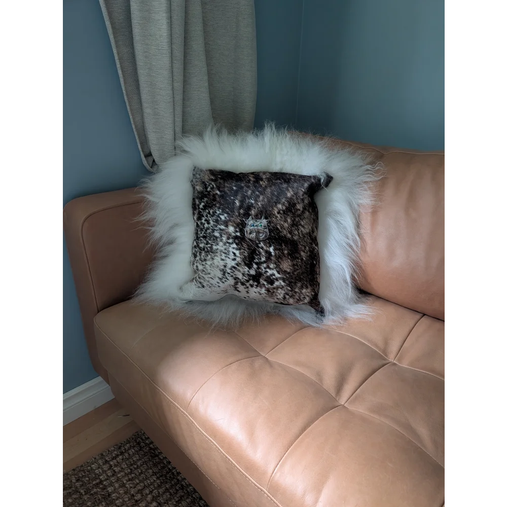 The moo cushion is a plush square pillow with a black and white speckled pattern and white faux fur trim on a tan couch