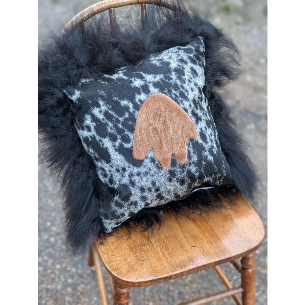 The yeti cushion cowhide features a black and white spotted pillow with a tan fuzzy ghost appliqué made from icelandic sheepskin and long wool