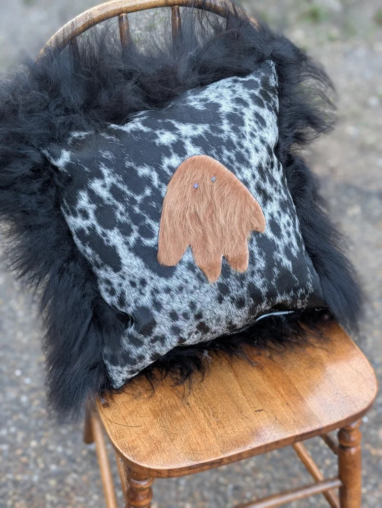 The yeti cushion cowhide features a black and white spotted pillow with a tan fuzzy ghost appliqué made from icelandic sheepskin and long wool