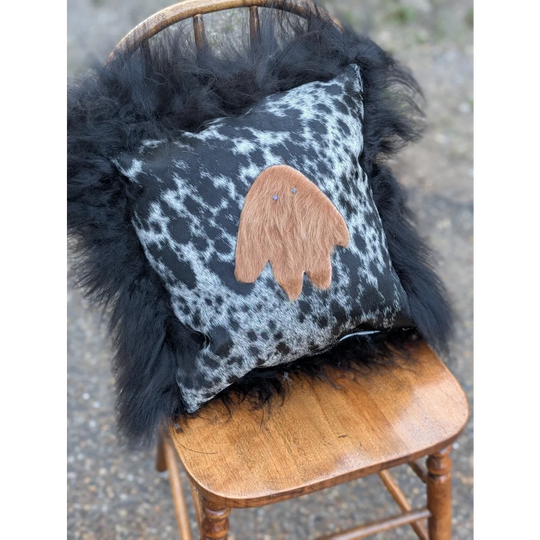 The yeti cushion cowhide features a black and white spotted pillow with a tan fuzzy ghost appliqué made from icelandic sheepskin and long wool