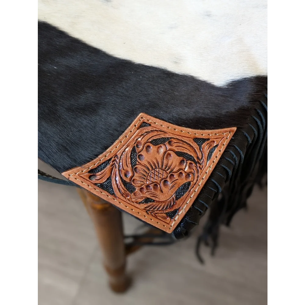 A hand tooled tan leather patch with floral design and black stitched edges on tumbleweed 2 tassel purse with zippered pocket