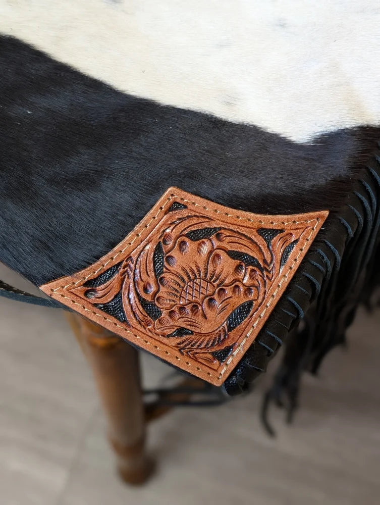 A hand tooled tan leather patch with floral design and black stitched edges on tumbleweed 2 tassel purse with zippered pocket