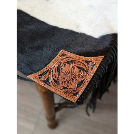 A hand tooled tan leather patch with floral design and black stitched edges on tumbleweed 2 tassel purse with zippered pocket
