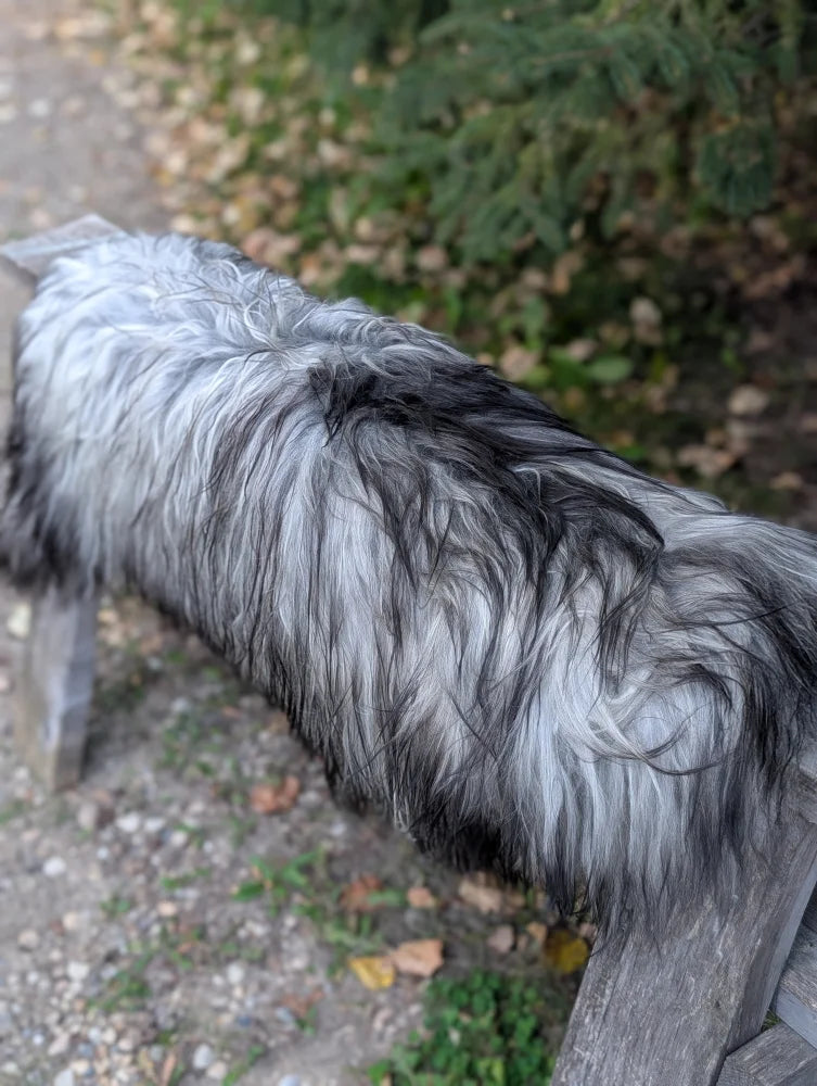 A long-haired dog stands on a gravel path next to a massive icelandic sheepskin rug in natural grey color