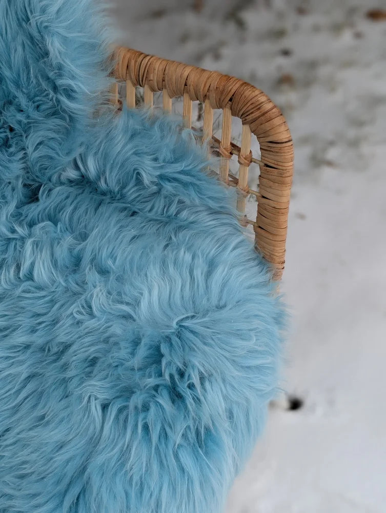 A plush light blue minty green merino sheepskin rug draped over a woven wicker chair armrest with soft fluffy texture