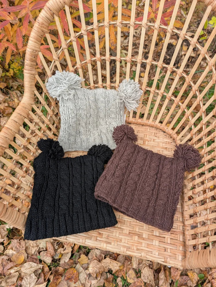 Three woolen heelah double pompom hats in gray black and brown with cable knit patterns and fluffy pompoms on top
